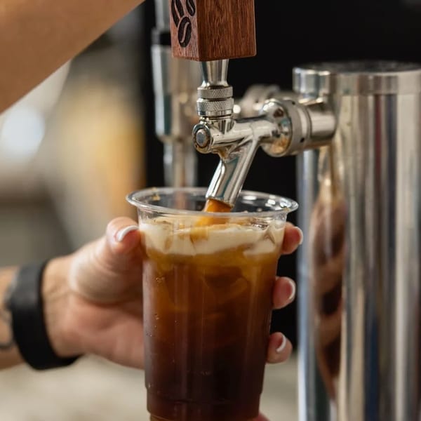 A barista pours a dark iced beverage with a creamy topping from a tap into a plastic cup.