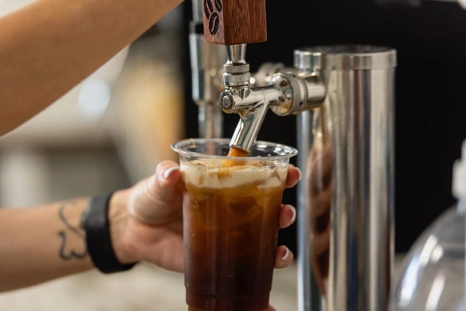 A barista pours a dark iced beverage with a creamy topping from a tap into a plastic cup.