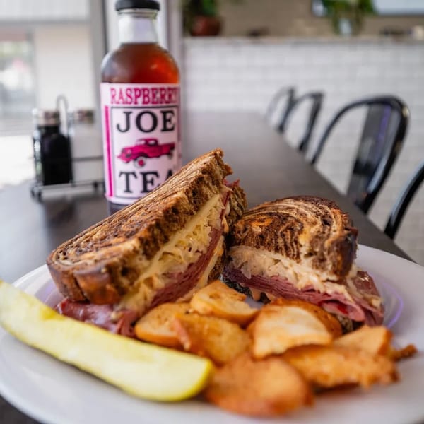 A Reuben sandwich, potato chips, and a pickle spear on a white plate with a bottle of Raspberry Joe Tea.