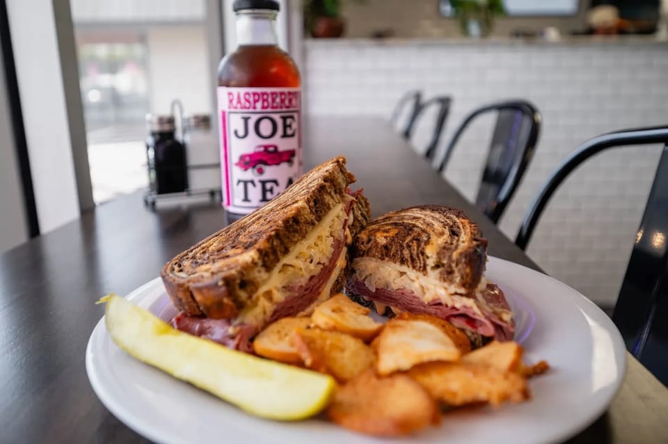A Reuben sandwich, potato chips, and a pickle spear on a white plate with a bottle of Raspberry Joe Tea.
