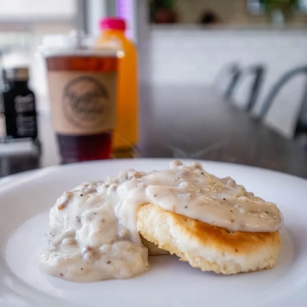 Two biscuits covered in creamy sausage gravy on a white plate, with drinks and chairs blurred in the background.