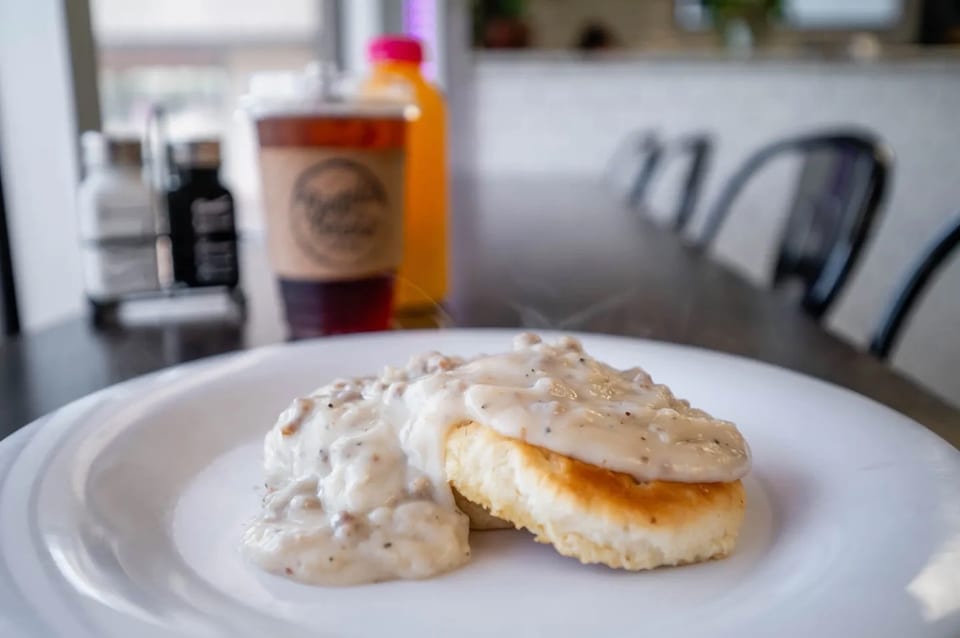 Two biscuits covered in creamy sausage gravy on a white plate, with drinks and chairs blurred in the background.