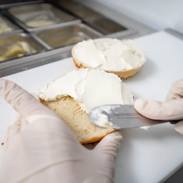 Gloved hands spreading cream cheese on a bagel half with a knife, another bagel half in the background.