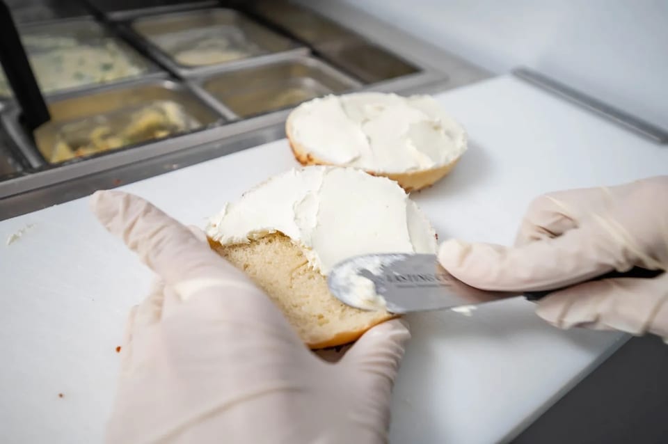 Gloved hands spreading cream cheese on a bagel half with a knife, another bagel half in the background.