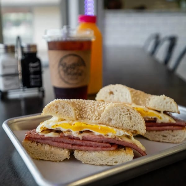 Two bagel sandwiches with fried eggs and meat, iced coffee, and orange juice on a table.