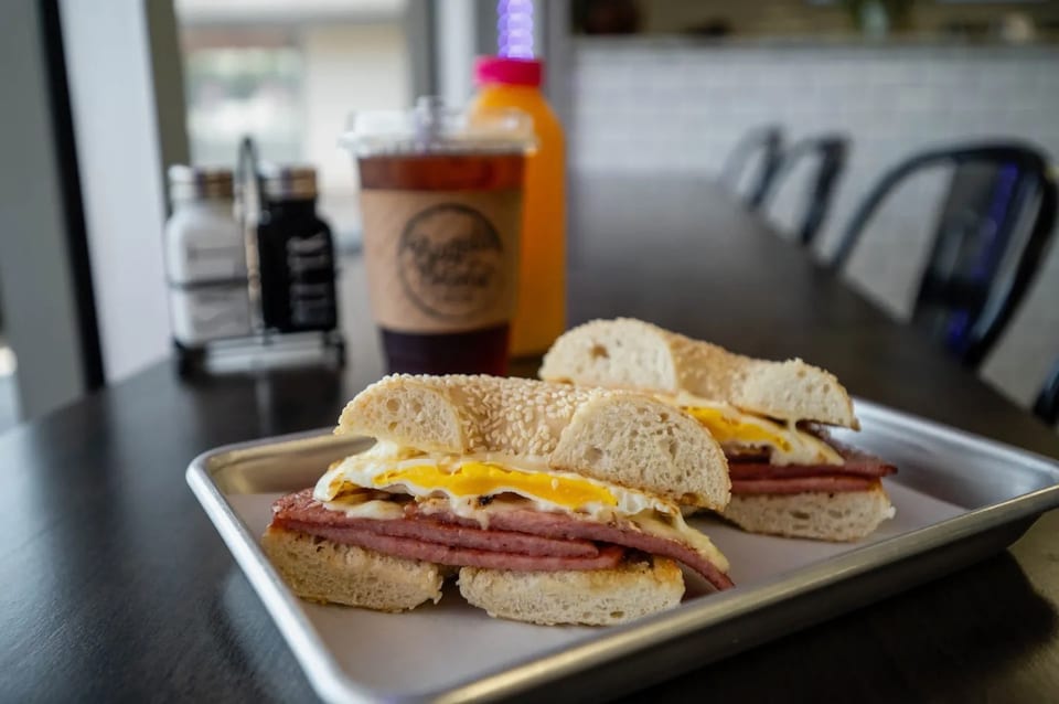 Two bagel sandwiches with fried eggs and meat, iced coffee, and orange juice on a table.